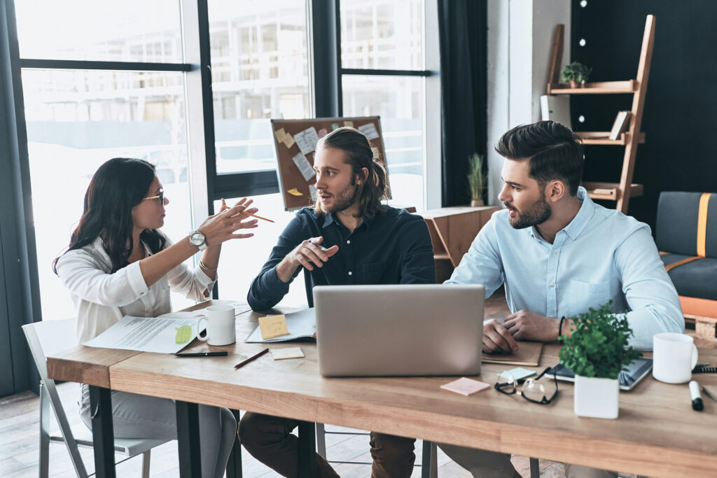 Equipe conversando de forma engajada ao redor de uma mesa de trabalho, com notebook e anotações, em um ambiente de escritório